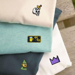 Pode incluir: Uma pilha de camisetas dobradas em creme, verde-azulado, verde-azulado escuro e branco. Cada camisa apresenta um design bordado pixelizado: uma caveira com uma espada, um copo de cerveja com texto, um gnomo e uma coroa roxa. As camisas estão empilhadas em uma superfície de madeira.