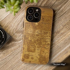 May include: A gold-toned phone case featuring a detailed map design with the text "Ancient Map." The map includes geographical features and place names such as "Wilderness," "Karamja," and "Kharidian Desert." The case is on a wooden surface.