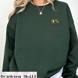 May include: A dark green sweatshirt with a small yellow and black graphic on the chest. The graphic appears to be a stylized beer mug. The text "Drinking Skill" is at the bottom of the image.