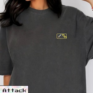 May include: Dark gray t-shirt with a small yellow and white graphic on the chest. The graphic features a sword and the numbers "99". The word "Attack" is displayed in a pixelated font at the bottom.