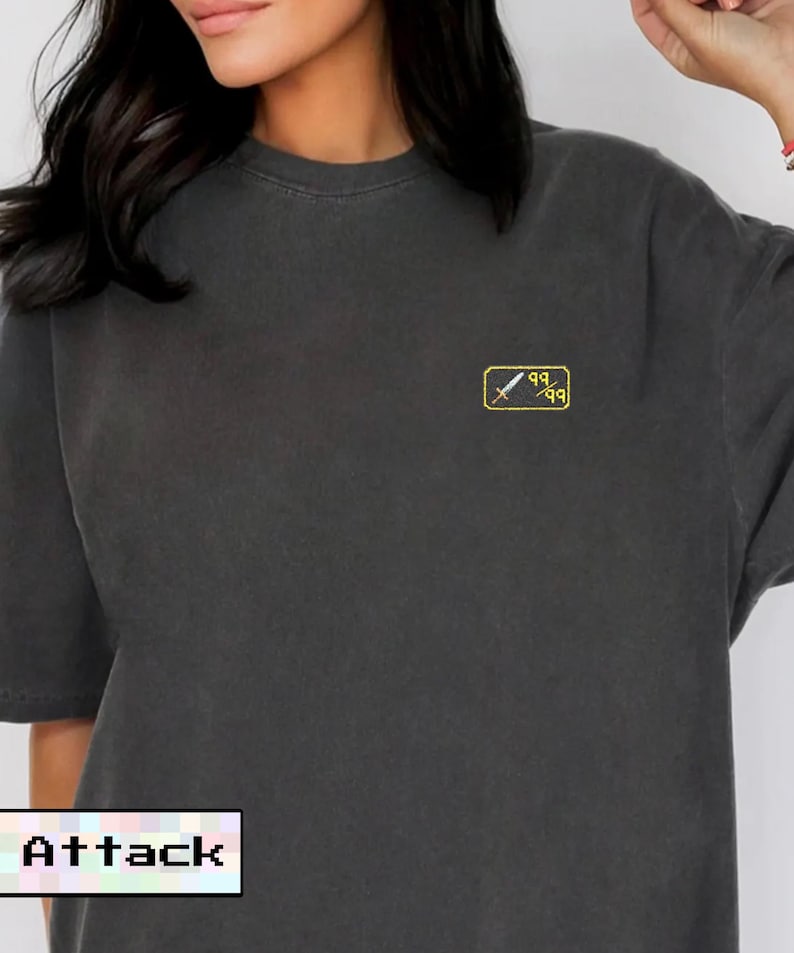May include: Dark gray t-shirt with a small yellow and white graphic on the chest. The graphic features a sword and the numbers "99". The word "Attack" is displayed in a pixelated font at the bottom.