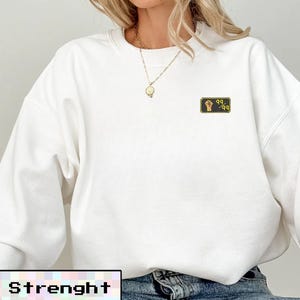 May include: White sweatshirt with a small embroidered patch featuring a fist and the numbers "99-99". The word "Strenght" is displayed in a pixelated font at the bottom of the image. The sweatshirt is paired with blue jeans.