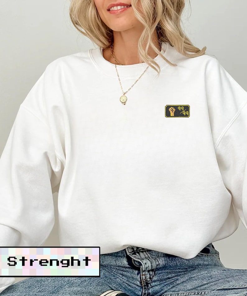 May include: White sweatshirt with a small embroidered patch featuring a fist and the numbers "99-99". The word "Strenght" is displayed in a pixelated font at the bottom of the image. The sweatshirt is paired with blue jeans.