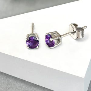 May include: A pair of stud earrings featuring round, deep purple gemstones set in silver-colored metal. The earrings are displayed on a white triangular surface, with a blurred floral arrangement in the background.
