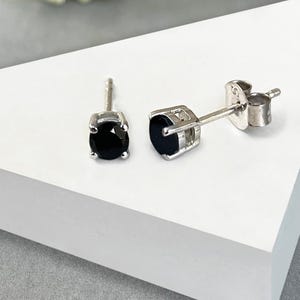 May include: A pair of stud earrings with black gemstones set in silver-colored metal. The earrings are displayed on a white geometric surface, with a soft-focus floral arrangement in the background.