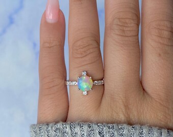 Pode incluir: Um anel de prata com uma grande pedra de opala redonda. A opala exibe um espectro de cores, incluindo azul, verde e amarelo. A banda é adornada com pequenas pedras claras. O anel é mostrado num dedo.