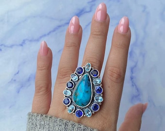 May include: A large silver ring featuring a teardrop-shaped turquoise stone surrounded by a halo of blue gemstones. The ring has a detailed, ornate design with various shades of blue stones.