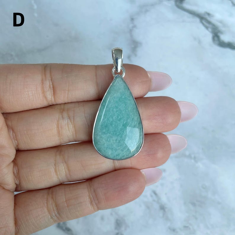May include: A teardrop-shaped pendant featuring a smooth, light green amazonite stone set in a polished silver-toned metal frame. The pendant includes a small silver-toned bail at the top for attachment to a chain or cord, showcasing a clean design.