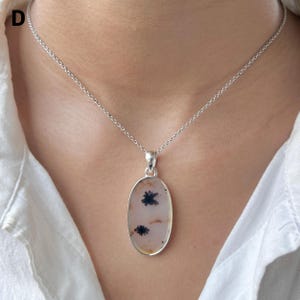 Puede incluir: Un collar de plata con un colgante ovalado de piedra lisa y transl&uacute;cida. La piedra es de color blanco roto con inclusiones dendr&iacute;ticas oscuras en forma de helecho repartidas por su superficie. El colgante cuelga de una fina cadena de plata.