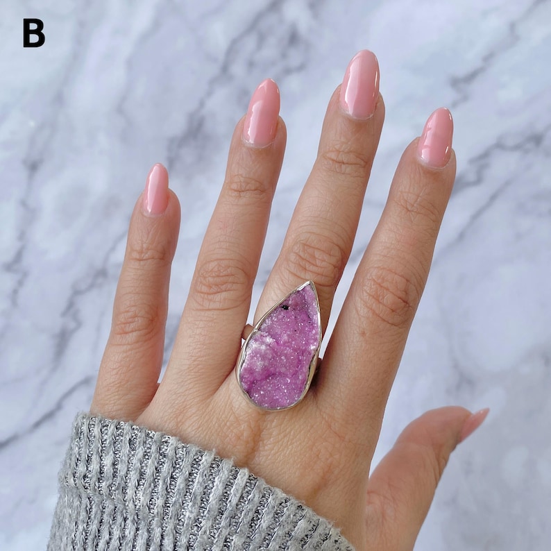 May include: A large, teardrop-shaped ring featuring a vibrant, textured pink gemstone set in a silver-colored metal band. The ring is displayed on a hand with pink nail polish, against a marble background.
