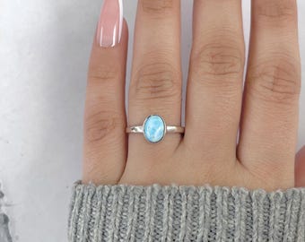 May include: A silver ring featuring an oval, light blue gemstone. The ring is set on a finger, showcasing the polished band and the vibrant color of the stone. The ring is a piece of jewelry.