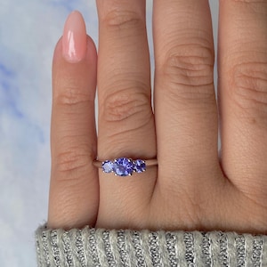 May include: A silver ring with three blue gemstones. The center stone is larger than the two flanking stones. The ring is displayed on a hand with a light pink nail polish and a gray sweater.