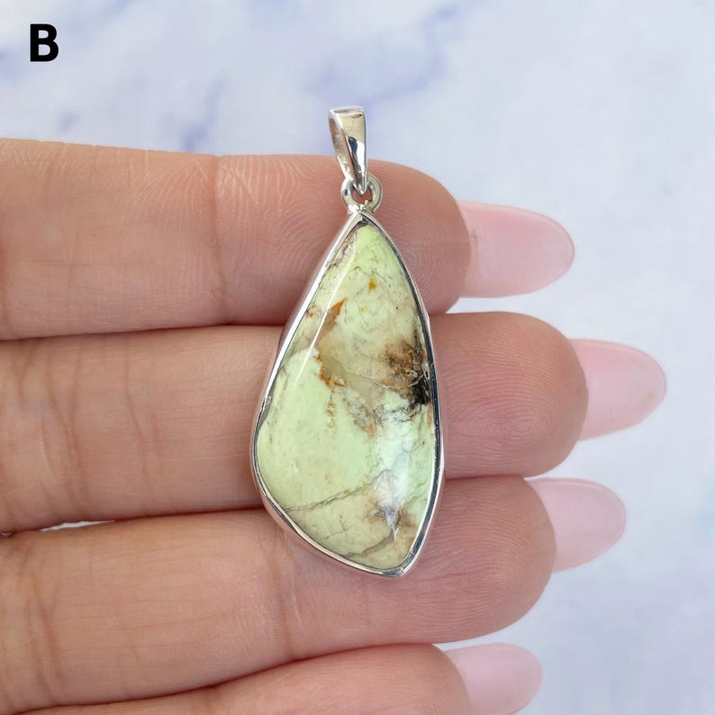 Pu&ograve; includere: Un ciondolo a forma di goccia con una pietra lucida verde pallido con venature marroni e nere, incastonato in una montatura in metallo argentato liscio con un piccolo gancio argentato sulla parte superiore, contrassegnato dalla lettera B.