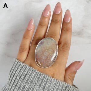 Pode incluir: Um anel oval com uma pedra grande e polida. A pedra tem um padrão creme e rosa, emoldurada por uma banda de metal prateado. O anel é exibido em uma mão, com um suéter cinza ao fundo.