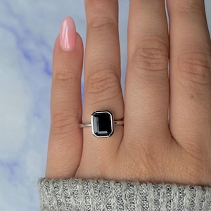 May include: A silver ring with a large, rectangular black gemstone. The gemstone is set in a bezel setting. The ring is displayed on a hand with pink nail polish. A classic and elegant piece of jewellery.
