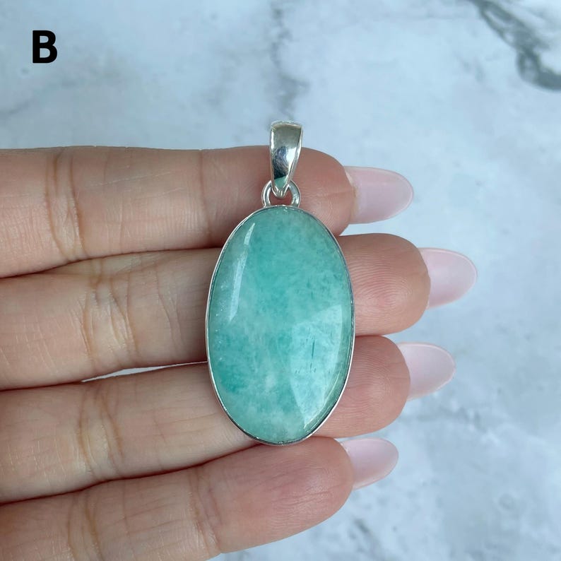 May include: An oval-shaped pendant featuring a smooth, polished light green amazonite gemstone set in a simple silver-toned metal frame with a small bail at the top for attachment to a chain. The stone displays natural variations in color.