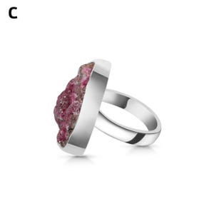 May include: A silver ring featuring a large, irregular-shaped, raw ruby gemstone. The ruby is a deep pinkish-red color with a textured surface. The ring band and setting are polished silver.