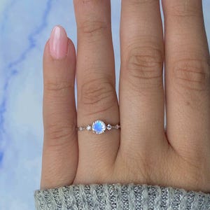 May include: A silver ring featuring a round, iridescent moonstone center stone, flanked by smaller, clear gemstones. The ring is set on a delicate band, showcasing a minimalist design. The ring is displayed on a hand with pink nail polish.