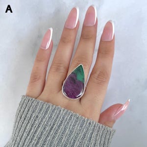 May include: A teardrop-shaped ring featuring a dual-toned stone with a deep purple base and a vibrant green top section, set in a polished silver-coloured metal band. The letter A is visible in the upper left corner of the frame.