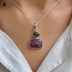 May include: A silver necklace featuring a pendant with a rectangular purple stone and a smaller, rounded green stone set above it. The pendant hangs from a thin silver chain, resting against a white fabric background, showcasing a metallic finish.