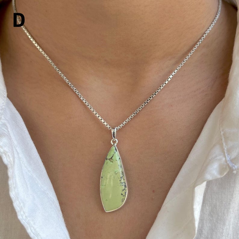 Pu&ograve; includere: Una collana a catena quadrata in argento con un ciondolo a goccia caratterizzato da una pietra liscia verde chiaro con motivi a matrice scuri e ramificati. La lettera D &egrave; visibile nell'angolo in alto a sinistra su una camicia bianca.