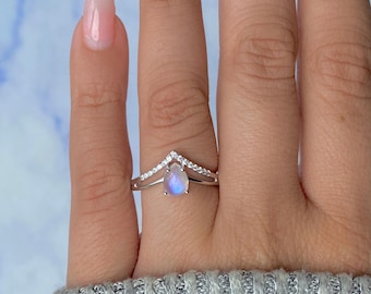 May include: A silver ring with a pear-shaped, iridescent moonstone. The ring features a chevron design with small, clear stones. The ring is worn on a finger, showcasing the jewelry's design and color.