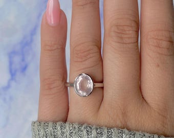 Puede incluir: Un anillo de plata con una gema ovalada de color rosa claro. El anillo tiene un engaste sencillo y la banda es fina y pulida. La gema es lisa y translúcida. El anillo se lleva en un dedo.