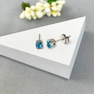Puede incluir: Un par de pendientes de botón con piedras preciosas redondas de color azul claro engastadas en metal plateado. Los pendientes se exhiben sobre una superficie geométrica blanca, con un arreglo floral borroso en el fondo. Una joya clásica.