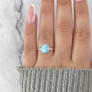 May include: A silver ring featuring an oval, light blue gemstone. The ring is set on a finger, with a gray sweater in the background. The gemstone has a marbled appearance, with shades of blue and white.