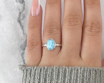 May include: A silver ring featuring an oval, light blue gemstone. The ring is set on a finger, with a gray sweater in the background. The gemstone has a marbled appearance, with shades of blue and white.