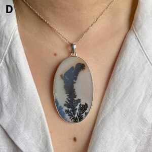 May include: An oval-shaped pendant necklace featuring a smooth, off-white stone with dark, tree-like dendritic inclusions. The stone is set in a simple silver-toned metal frame and hangs from a delicate, matching silver-toned chain link necklace.