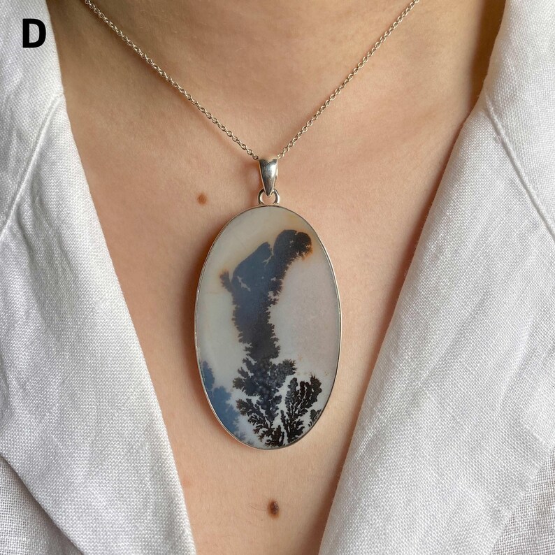 May include: An oval-shaped pendant necklace featuring a smooth, off-white stone with dark, tree-like dendritic inclusions. The stone is set in a simple silver-toned metal frame and hangs from a delicate, matching silver-toned chain link necklace.