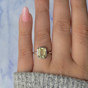 May include: A silver ring featuring a rectangular, pale yellow gemstone. The gemstone is set within a bezel. The ring is on a finger with pink nail polish.