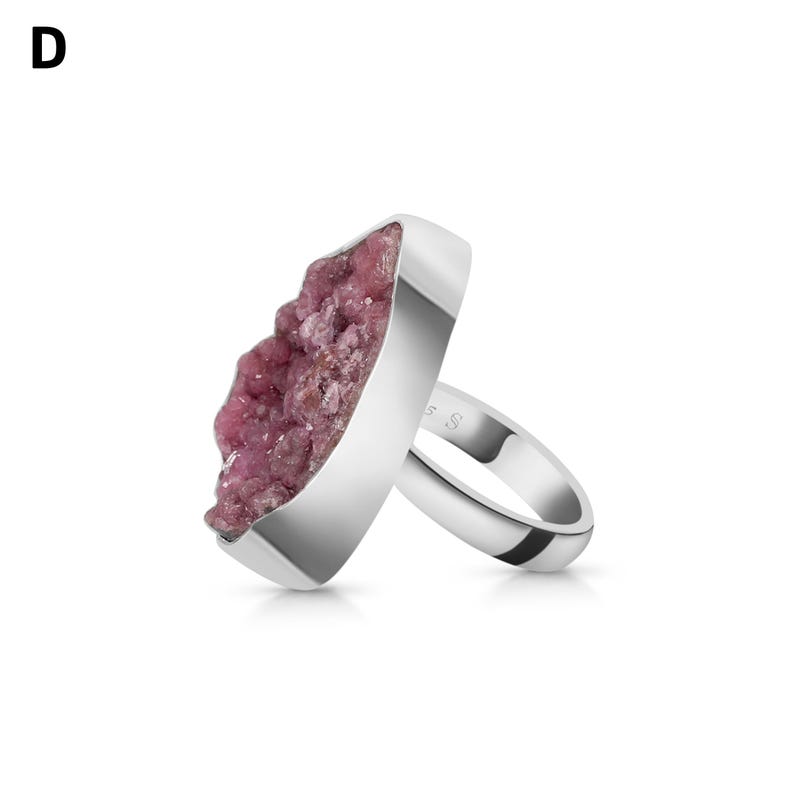 May include: A silver ring featuring a large, irregular-shaped pink gemstone. The gemstone has a textured, crystalline appearance. The ring's band is smooth and polished, with a minimalist design. The ring is set against a white background.
