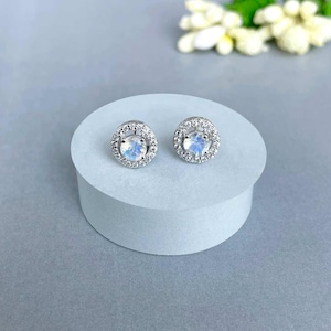 May include: A pair of silver stud earrings with a central, round, iridescent stone surrounded by a halo of smaller, clear stones. The earrings are displayed on a white, cylindrical pedestal against a soft gray background.