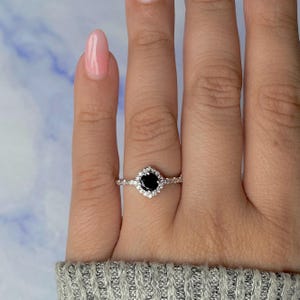 Può includere: Un anello d'argento con una pietra nera centrale e un alone di pietre bianche più piccole. L'anello è indossato su una mano con smalto rosa, su uno sfondo blu e bianco.
