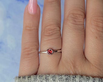 Op de afbeelding: Een zilveren ring met een ronde, rode edelsteen. De ring heeft een smalle band. De edelsteen is gezet in een bezel setting. De ring wordt getoond op een vinger. Eenvoudig en elegant ontwerp.