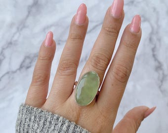 May include: A silver ring featuring a large, oval-shaped, pale green gemstone. The ring's setting is a simple, polished silver band. The gemstone has a translucent quality, and the ring is worn on a hand.