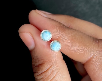 May include: A pair of round stud earrings featuring polished light blue stones with white marbling, each set in a simple silver-toned metal bezel. The circular stones have a smooth, domed surface and are held together in a small, compact arrangement.
