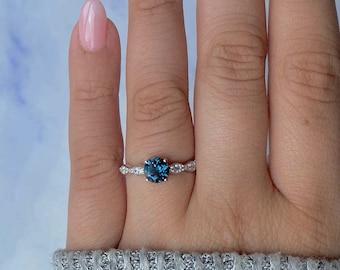 May include: A silver ring with a large, round, deep blue gemstone as the centerpiece. The band is adorned with smaller, clear gemstones. The ring is set against a neutral background, highlighting its elegant design. A piece of jewelry.