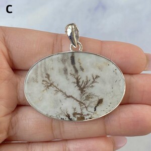 May include: An oval-shaped pendant featuring a white stone with dark brown, tree-like dendritic inclusions. The stone is set in a polished silver-colored metal frame with a matching silver-colored bail at the top for hanging on a chain.