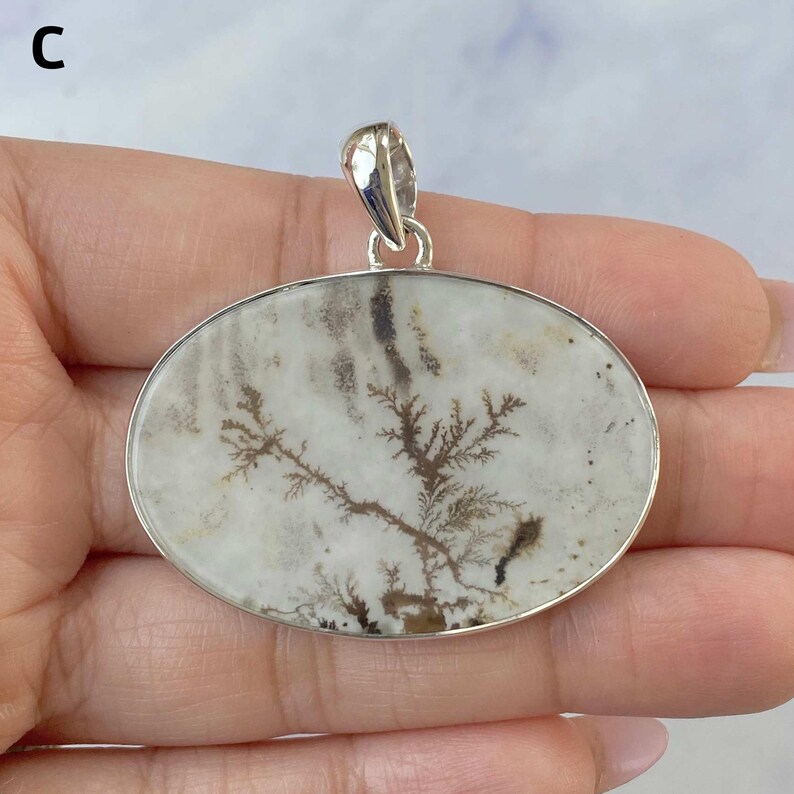 May include: An oval-shaped pendant featuring a white stone with dark brown, tree-like dendritic inclusions. The stone is set in a polished silver-colored metal frame with a matching silver-colored bail at the top for hanging on a chain.