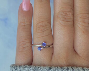 May include: A silver ring with two round, blue gemstones. The ring has a delicate, curved design, with the gemstones set on either side of the finger. The ring is shown on a person's finger, with a light blue and white background.