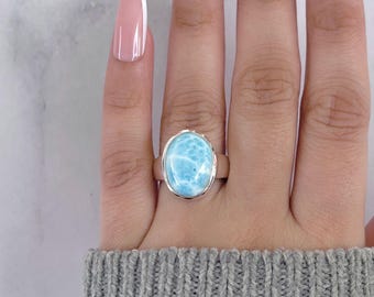 May include: A silver ring featuring a large, oval-shaped, light blue gemstone. The gemstone has a marbled appearance with white veining. The ring is set on a silver band and is displayed on a hand.
