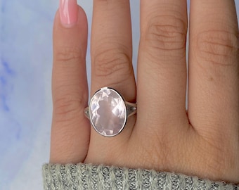 Puede incluir: Un anillo de plata con una gran piedra de cuarzo rosa de corte ovalado. La piedra rosa pálido está engastada en una banda de plata pulida. El anillo se muestra en una mano con esmalte de uñas rosa claro.