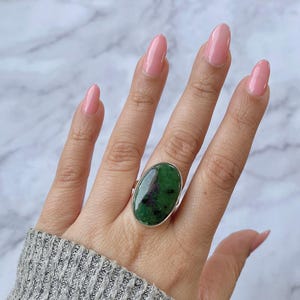 May include: A silver ring featuring a large, oval-shaped green gemstone with dark red inclusions. The ring is set on a hand with pink nail polish, against a marble background. The gemstone is the focal point of the jewelry.