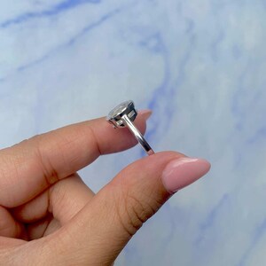 May include: A silver ring with a large, oval-shaped, clear gemstone. The ring is held up against a blue and white background. The ring's band is thin and polished.