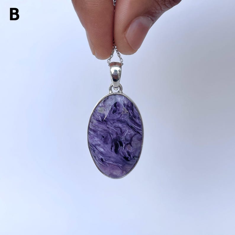 Op de afbeelding: Een ovaalvormige hanger met een gepolijste paars en zwart gemarmerde steen in een zilverkleurige metalen zetting. De hanger hangt aan een fijne zilverkleurige ketting. De letter B is zichtbaar in de linkerbovenhoek van de afbeelding.