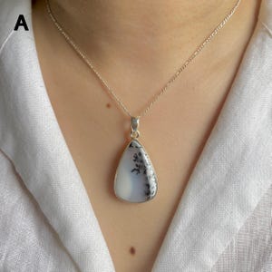 May include: A silver necklace featuring a teardrop-shaped pendant with a smooth, milky white stone containing dark, fern-like dendritic inclusions. The pendant is suspended from a delicate silver chain with a classic link design.