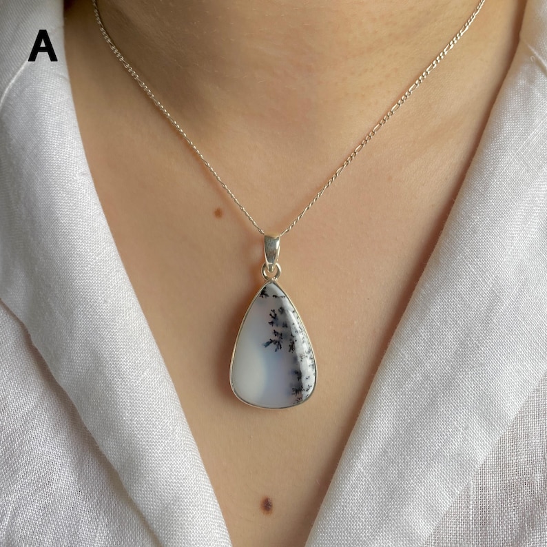 May include: A silver necklace featuring a teardrop-shaped pendant with a smooth, milky white stone containing dark, fern-like dendritic inclusions. The pendant is suspended from a delicate silver chain with a classic link design.